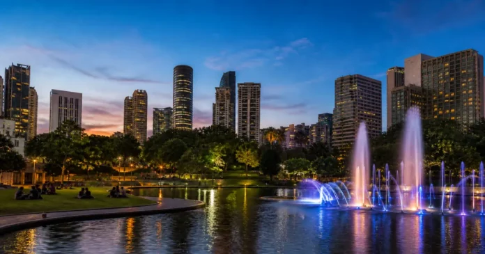 City park at dusk with a lit fountain and illuminated skyline behind